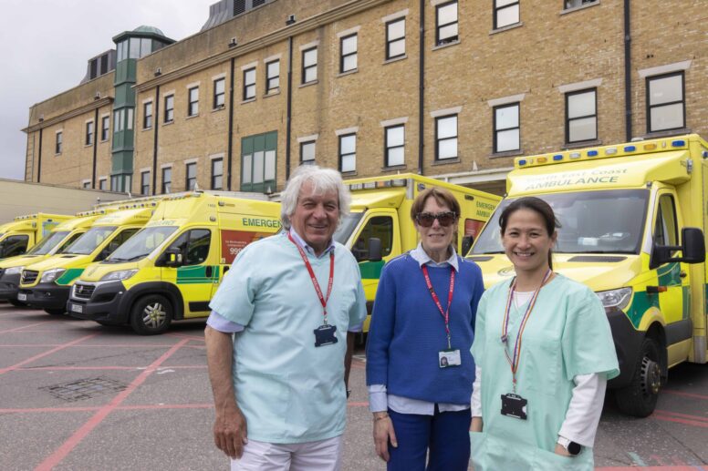 3 volunteers standing in front of parked ambulances outside a hospital building.